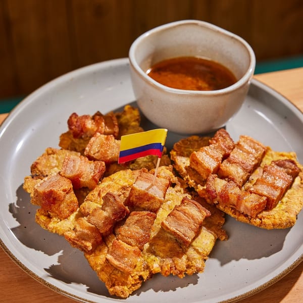 Colombian tostones topped with crispy pork belly and a small Colombian flag, served with a dipping sauce.