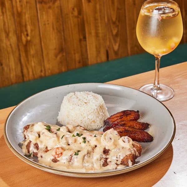 A Colombian dish with meat in creamy sauce, rice, fried plantains, and a drink on a wooden table.