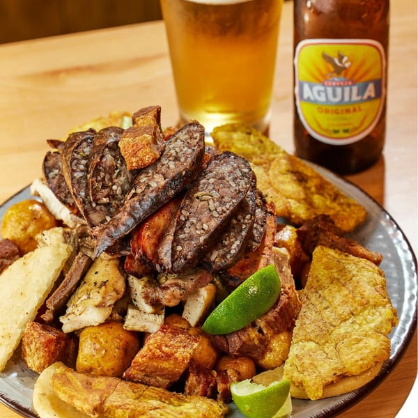 A large platter of Colombian picada with morcilla, chicharron, potatoes, and plantains, with an Aguila beer.