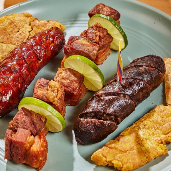 Colombian platter with chorizo, morcilla, chicharrón, and patacones, garnished with lime and a Colombian flag.