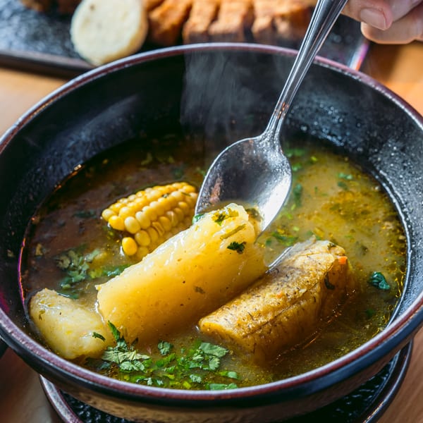 Steaming Colombian sancocho soup with corn, plantain, and yucca, being served with a spoon.