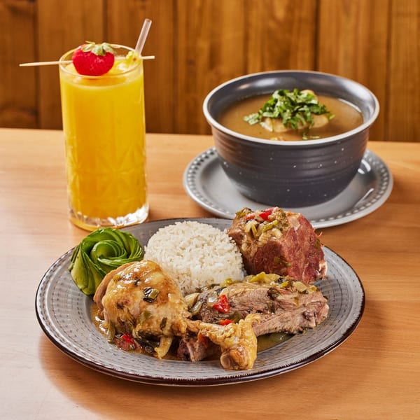 Colombian meal with chicken, beef, rice, avocado, soup, and a strawberry-garnished drink on a wooden table.