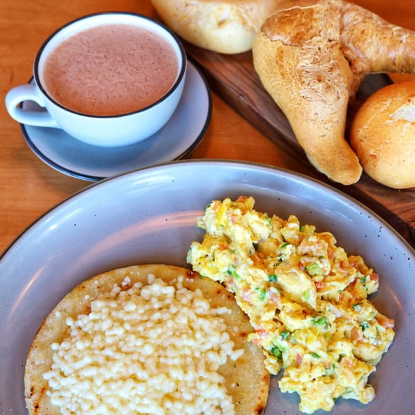 Colombian breakfast with scrambled eggs, cheesy arepa, hot chocolate, and various breads on a wooden table.