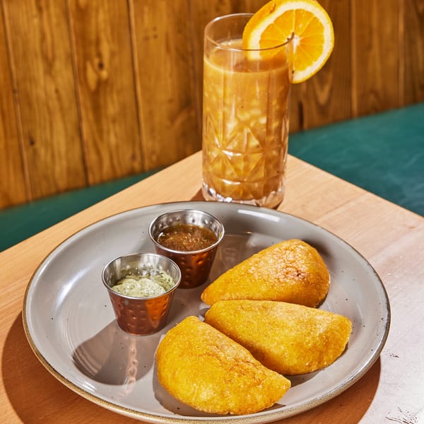 Three golden-fried Colombian empanadas with two dipping sauces and an orange juice on a wooden table.